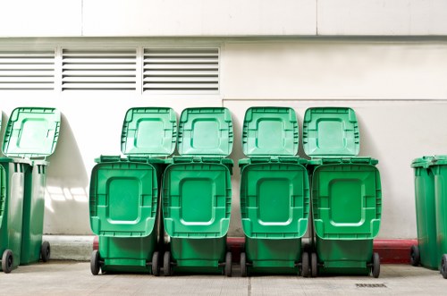Six green wheelie bins with closed lids are lined up against a white exterior wall, which features horizontal vent slats near the top. The bins are positioned on a paved surface adjacent to a building in a residential or commercial area, with some bins slightly leaning against each other. The image captures the bins from a straight-on angle, highlighting the uniformity of their design, with black wheels visible at the base. A narrow red line runs along the base of the building wall behind the bins. This scene is representative of waste collection or rubbish management in South Kensington, demonstrating the typical equipment used for rubbish removal services by companies such as Garden Clearance South Kensington, which operates in areas like SW7 postcode district. The lighting suggests daylight, with soft shadows cast on the ground, emphasizing a clean and organized environment suitable for waste removal activities.