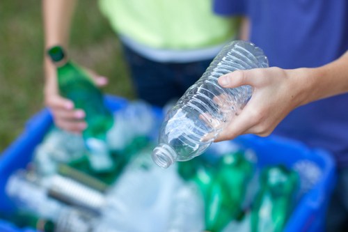 A close-up of a person's hand holding a clear plastic bottle with a slightly tapered neck and ridges near the top, ready for disposal or recycling. In the background, two individuals are seen, one wearing a yellow vest and the other in a blue shirt, standing near a blue plastic recycling bin filled with various green plastic bottles and other plastic waste. The setting appears to be outdoors on a grassy area, with the focus primarily on the plastic bottle in the foreground. The scene illustrates waste collection in progress, aligning with rubbish removal services offered by Garden Clearance South Kensington for local garden, household, or commercial waste disposal.