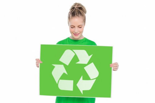 A young woman with blonde hair tied back in a ponytail, wearing a bright green T-shirt, holds up a large green sign featuring a white recycling symbol composed of three chasing arrows forming a triangle. She is smiling and looking down at the sign, standing against a plain white background. The image emphasizes environmental consciousness and recycling awareness, which relates to rubbish removal services offered by Garden Clearance South Kensington. The simplicity of the background and the clear focus on the woman and the recycling symbol make the visual suitable for conveying eco-friendly messages associated with local rubbish collection and waste management initiatives in South Kensington and surrounding areas. The image of the recycling sign symbolizes sustainable waste disposal and environmental responsibility, aligning subtly with the company's category of services handling rubbish clearance.