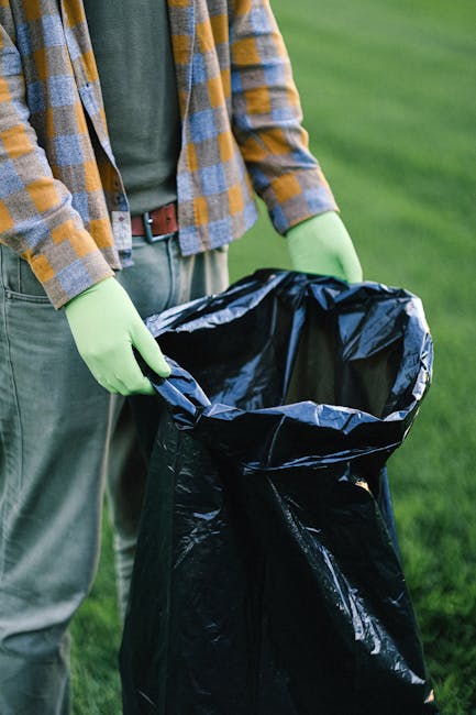 A person wearing a plaid yellow and gray jacket over a green shirt, with olive-green gloves, is holding open a black large plastic rubbish bag with metal tie handles. The background shows a well-maint