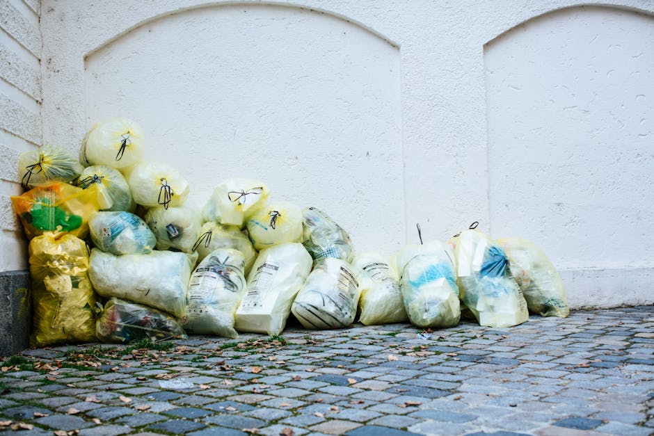 A person wearing a plaid yellow and gray jacket over a green shirt, with olive-green gloves, is holding open a black large plastic rubbish bag with metal tie handles. The background shows a well-maint