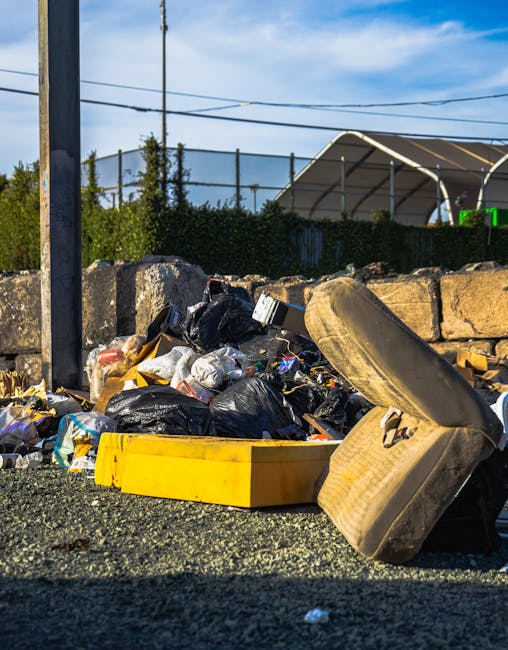 A pile of discarded cardboard boxes, some flattened and others partially intact, alongside plastic and cloth wrapping, is seen in a corner next to a brick and concrete wall. The debris includes packag