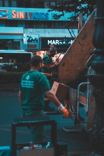 Two waste collection workers are seen outdoors at dusk, engaged in loading or unloading rubbish materials into a refuse vehicle. The worker in the foreground, with short hair, wears a teal-green unifo