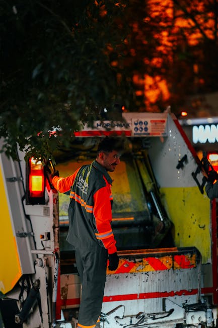 Two waste collection workers are seen outdoors at dusk, engaged in loading or unloading rubbish materials into a refuse vehicle. The worker in the foreground, with short hair, wears a teal-green unifo